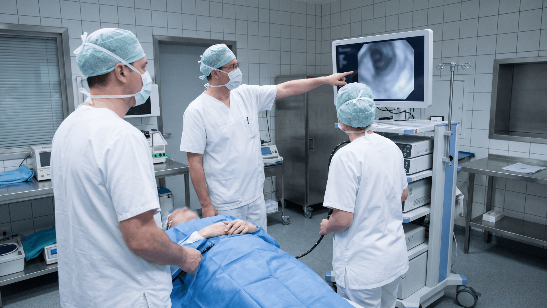 Drei medizinische Fachkräfte in Schutzkleidung betrachten bei einer endoskopischen Untersuchung eine Aufnahme auf dem Monitor. Eine Person liegt auf der Untersuchungsliege.