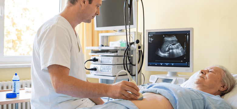 Medizinische Fachkraft führt eine Ultraschalluntersuchung des Bauchraums bei einer liegenden Patientin durch. Auf dem Monitor ist eine Ultraschallaufnahme zu sehen.