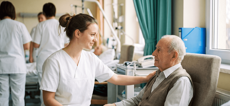Junge Frau in weißer Pflegekleidung betreut einen älteren Patienten im Krankenhauszimmer, legt ihm eine Hand auf die Schulter und hält ein Klemmbrett mit Unterlagen.