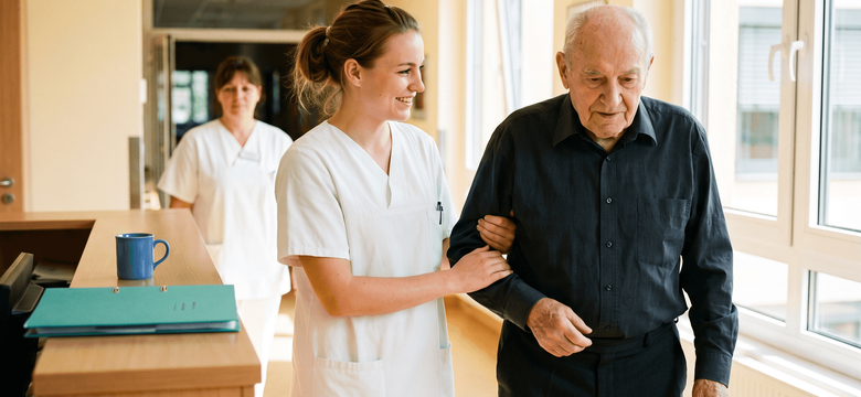 Junge Frau in weißer Pflegekleidung unterstützt einen älteren Patienten beim Gehen durch einen hellen Krankenhausflur und hält ihn dabei am Arm.