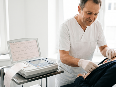 Medizinische Fachkraft in weißer Arbeitskleidung und Einmalhandschuhen bringt EKG-Elektroden am Brustkorb eines liegenden Patienten an. Daneben steht ein EKG-Gerät mit aufgezeichneten Herzstromkurven.